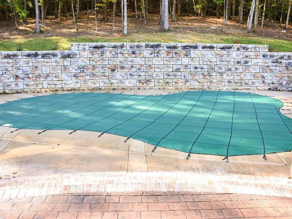 A green pool cover is sitting on a brick patio next to a stone wall.
