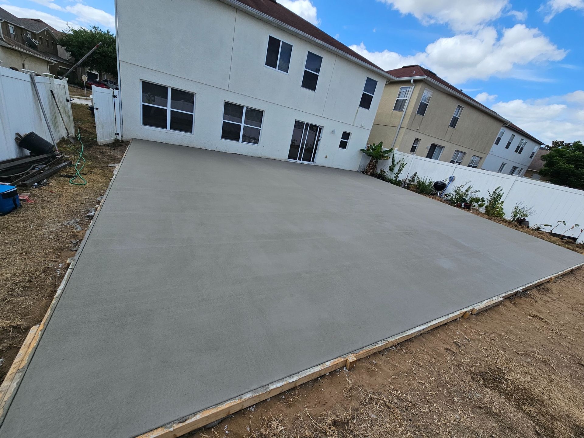 Newly poured concrete patio in a residential backyard, next to a white fence and houses.