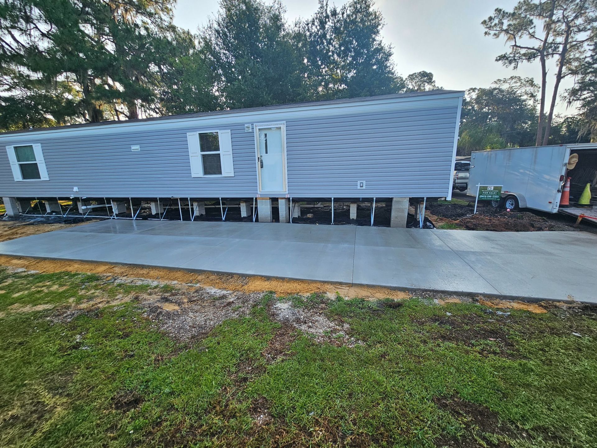 Mobile home on concrete pad; gray siding, white door, and supports. Green grass in the foreground.
