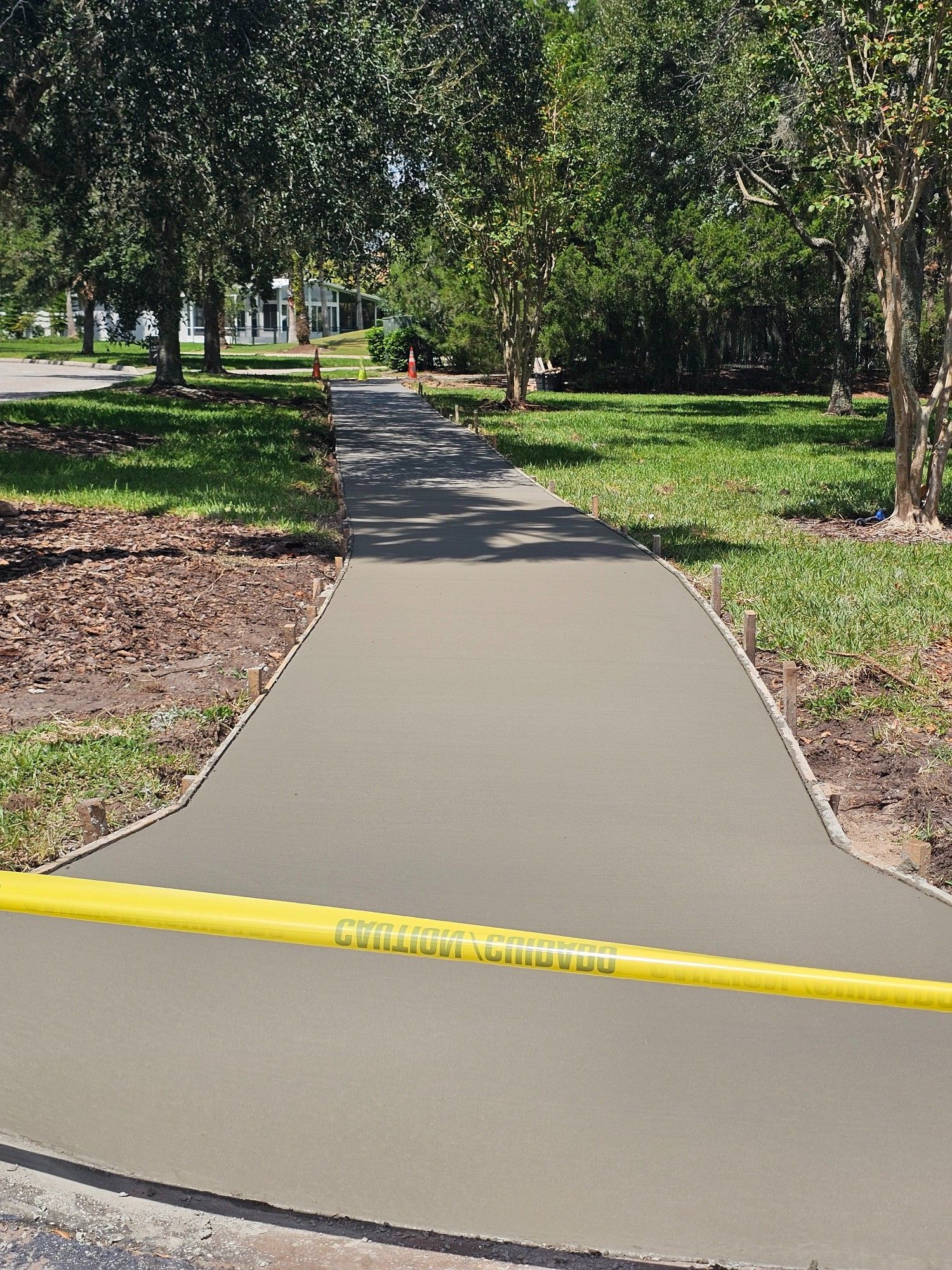 Newly poured gray concrete sidewalk, yellow caution tape, trees in the background, sunny day.
