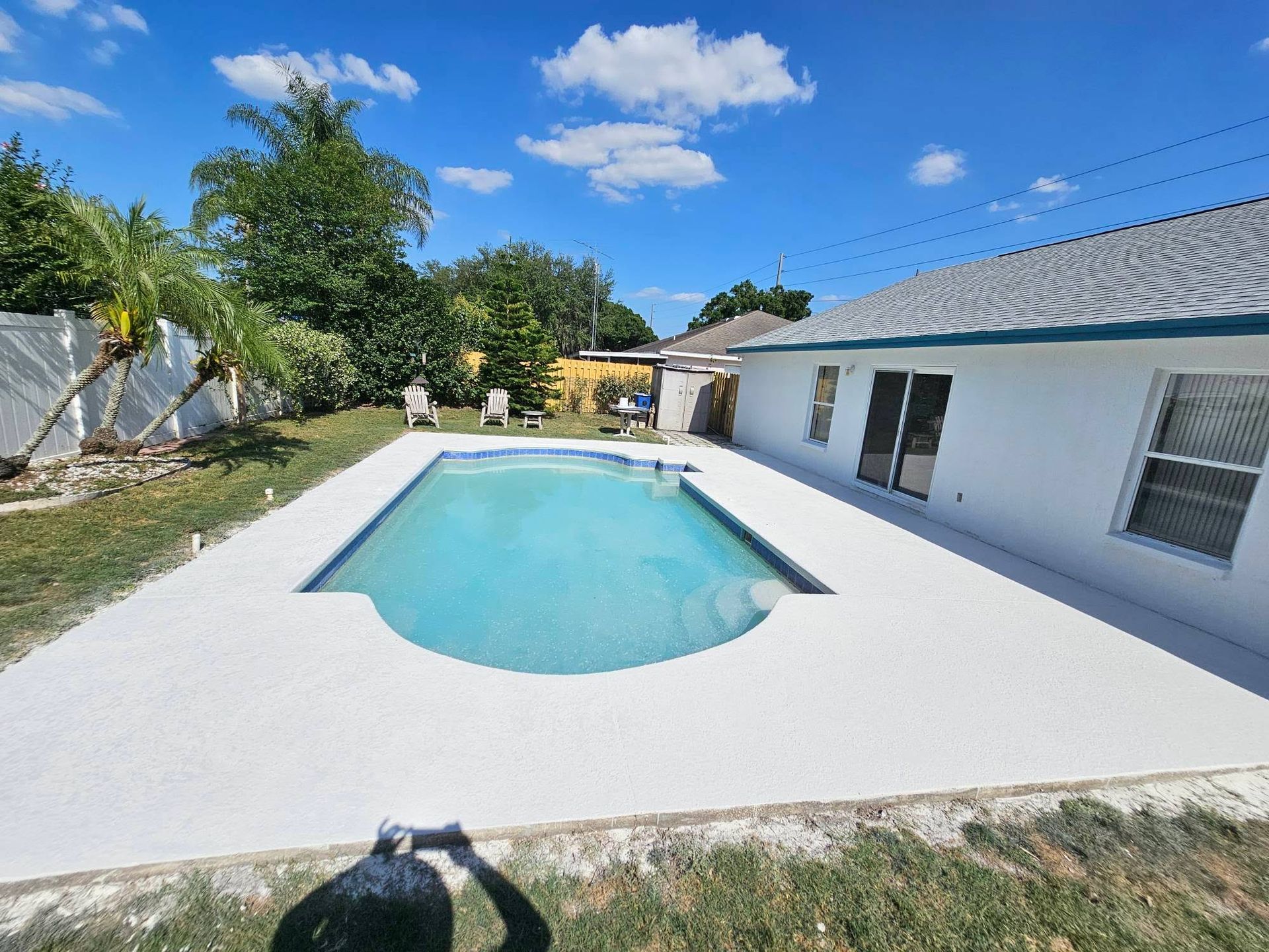 Backyard with pool, white concrete deck, and a white house under a blue sky with fluffy clouds.