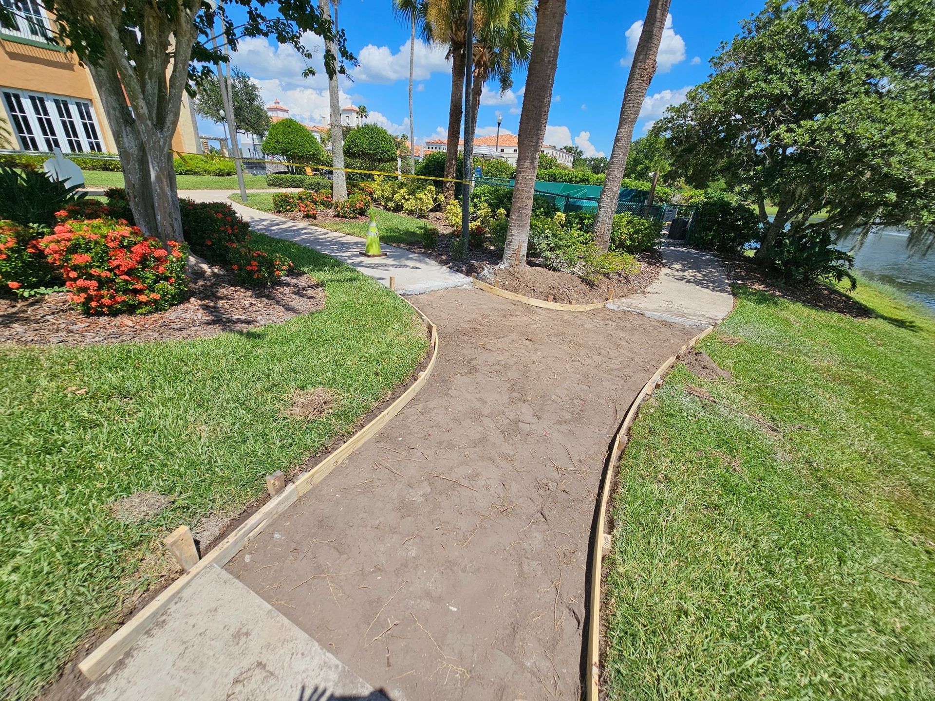 Pathways in a park with grass, trees, and flowers. One path is paved, the other dirt, leading to a lake.