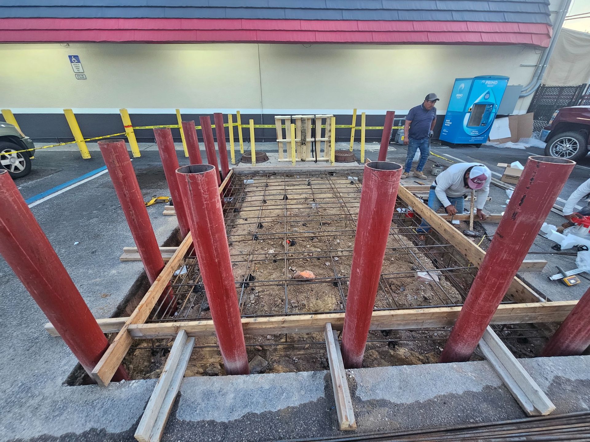 Construction site with red posts, wooden frames, and workers in a parking lot.