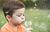 Boy blowing dandelion seeds in a green field; he wears an orange shirt and has closed eyes.