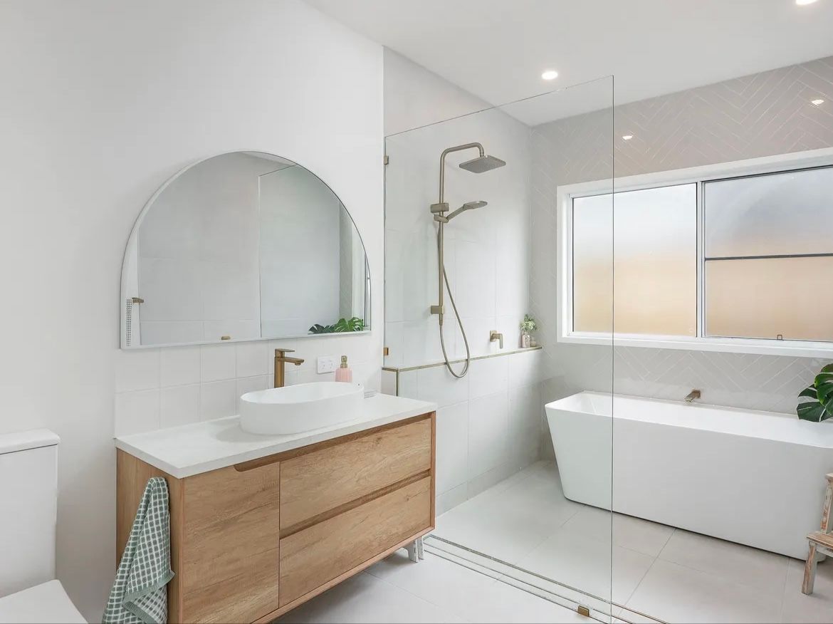 Modern Bathroom With Wooden Vanity, Arched Mirror, Glass Shower, And A White Tub — JGD Constructions in Atherton, QLD