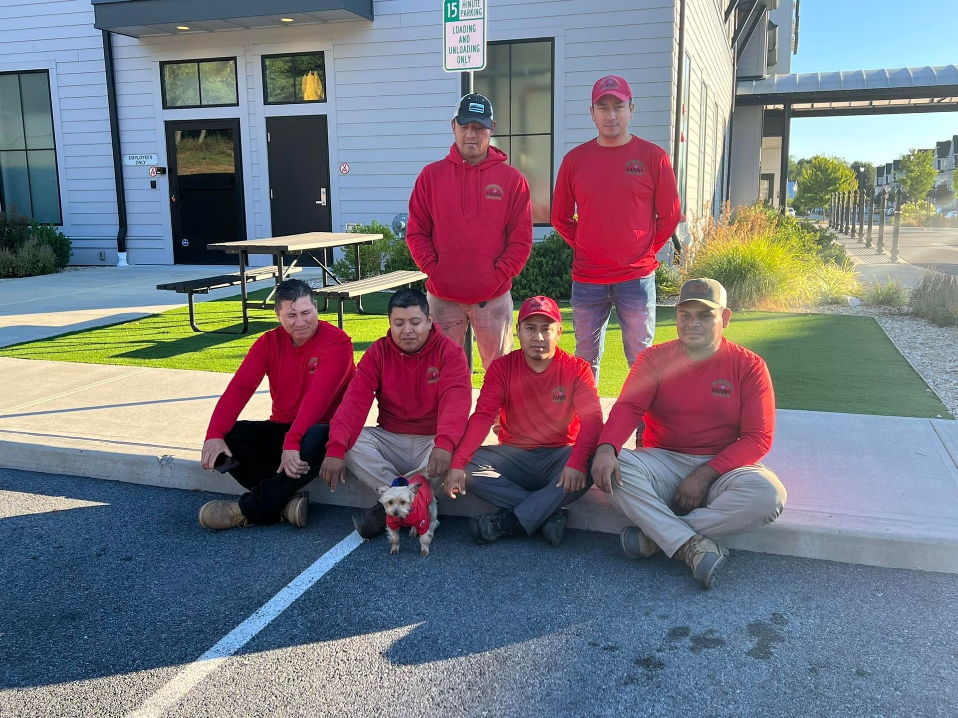 Six people in red shirts and a small dog pose outdoors by a building.