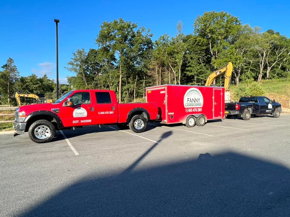 Red truck towing a red trailer and a black truck in a parking lot, with trees and construction equipment in the background.
