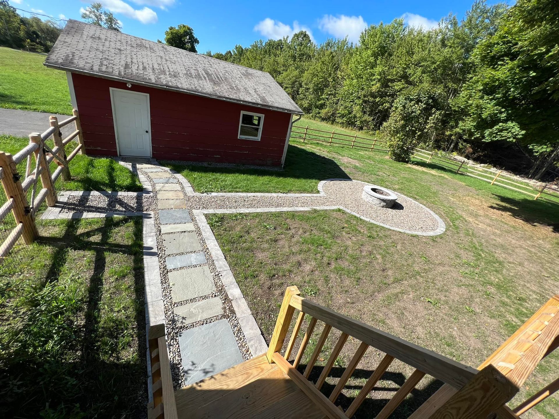 Red shed with stone path and fire pit on a grassy hill. Wooden fence and stairs. Sunny day.