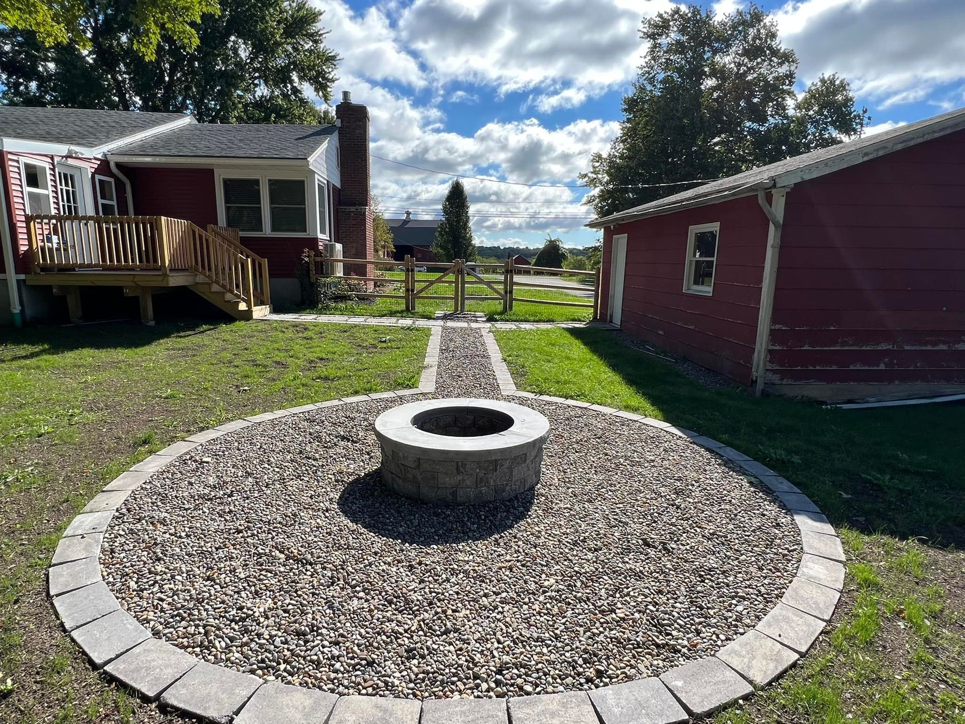 Backyard with fire pit, gravel, and stone path leading to a red building. Sunny day.
