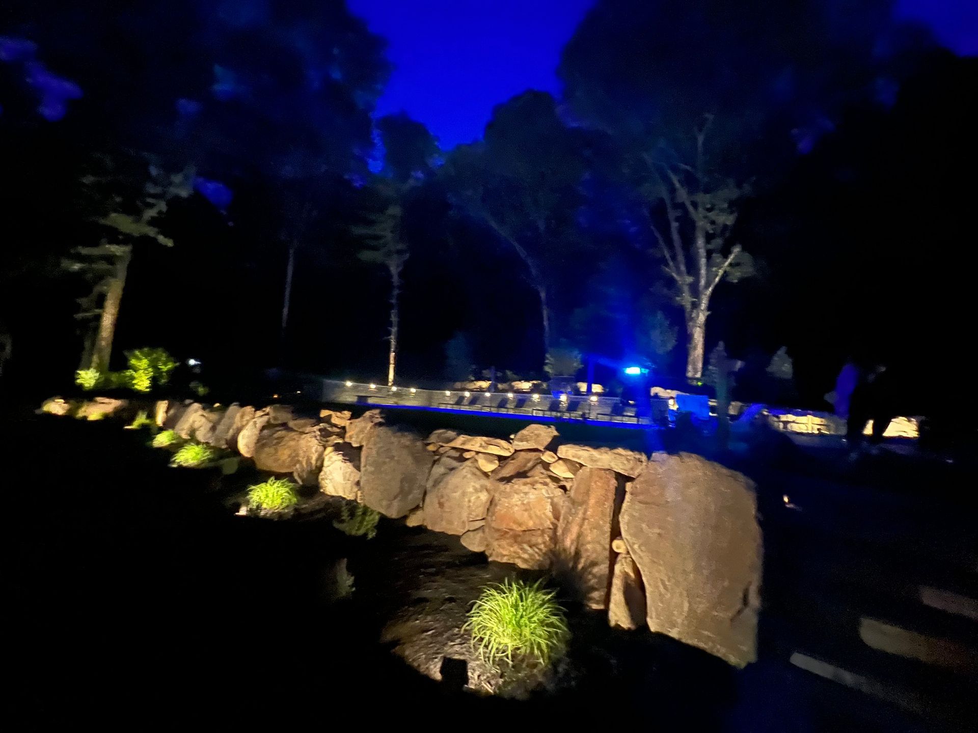 Night landscape with stone wall, lit plants, and trees under a blue sky.