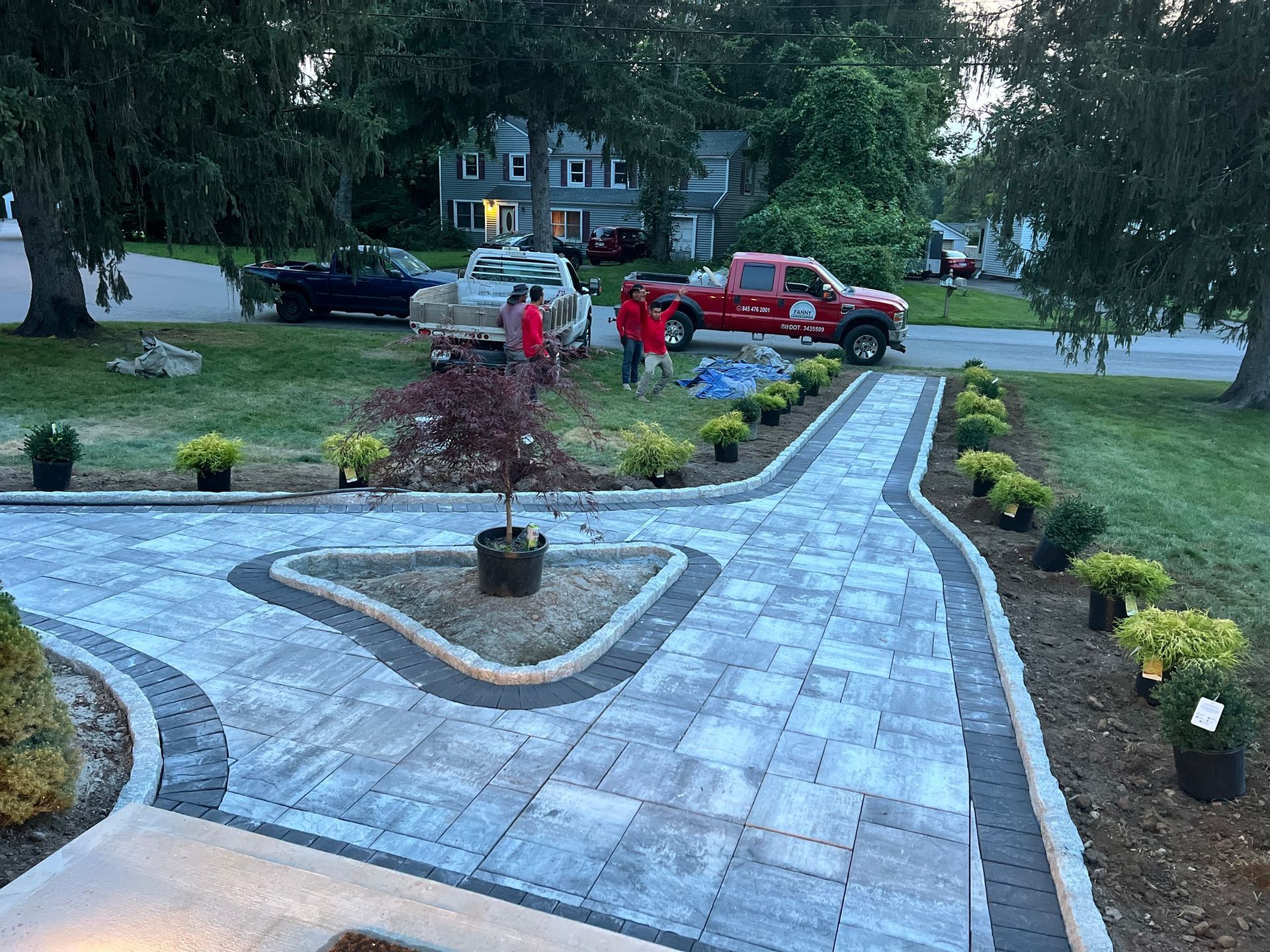 Driveway with pavers, lined with greenery, red trucks in background, workers preparing the area.