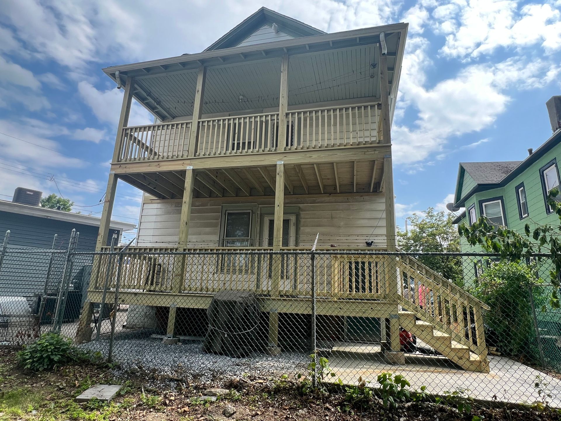 Back of a weathered two-story house with wooden decks and stairs, viewed from a yard with a chain-link fence.