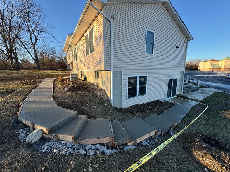Newly poured concrete walkway and steps next to a two-story house; construction in progress, safety tape present.