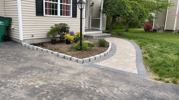 Paved walkway with flower beds and bordering stones leading to a beige house, with a black lamp post.