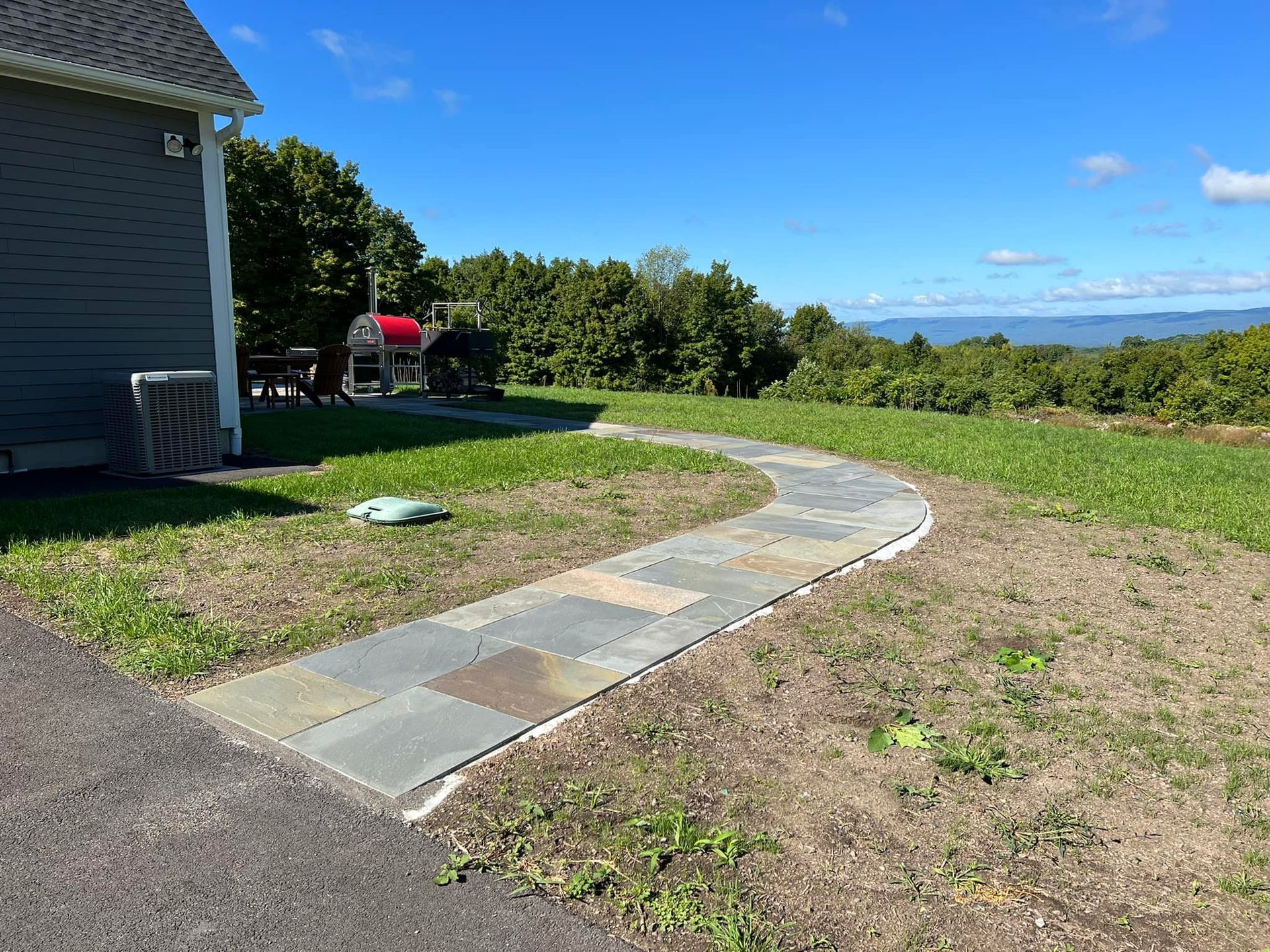 Stone path curves through grass, leading to a red grill near a house. Clear sky, trees, and hills in background.