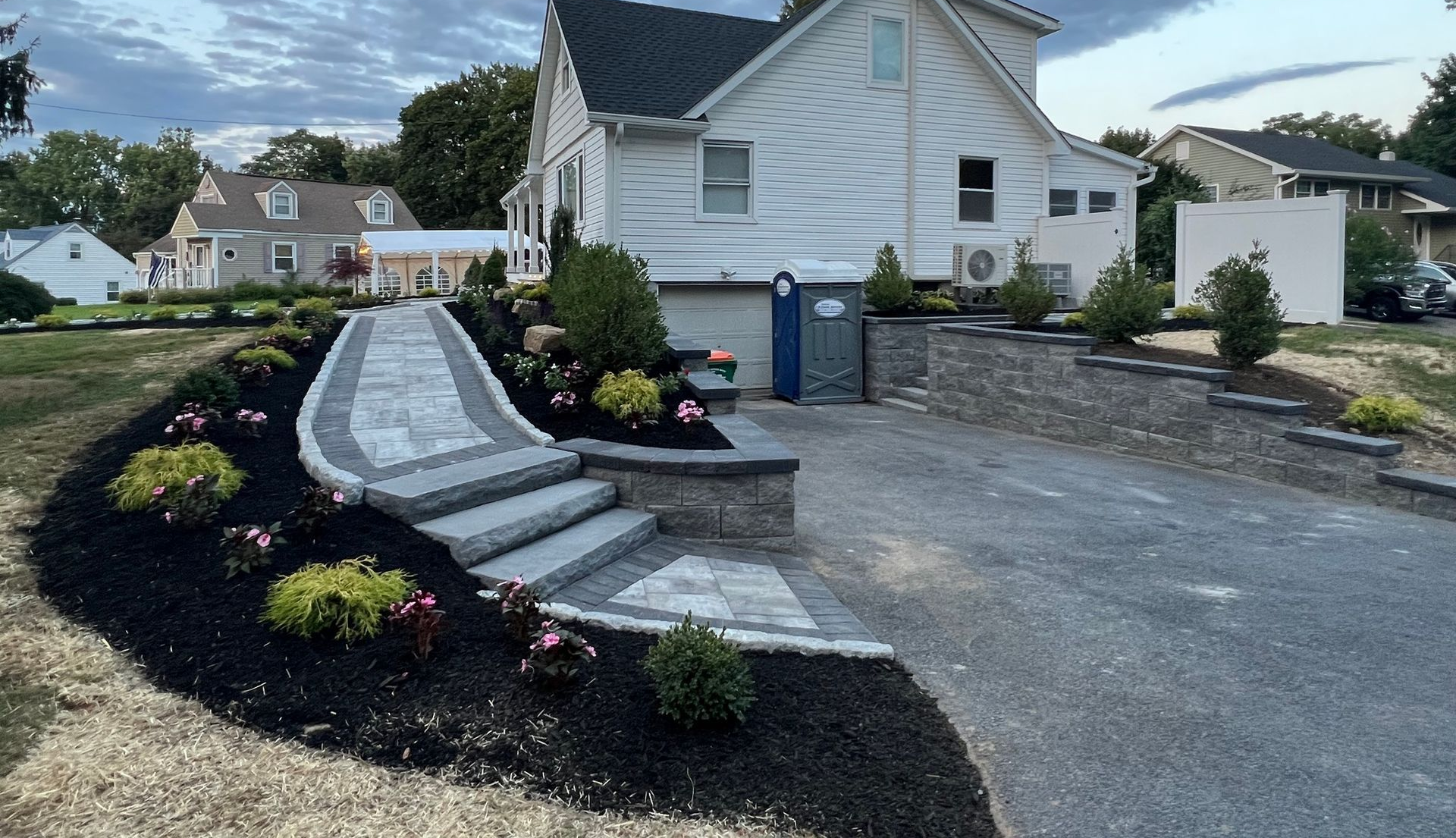 Pathway with steps leads to a white house with landscaped beds, dark mulch, and a paved driveway.