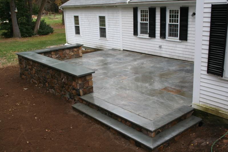 Stone patio with steps and low retaining wall next to a white house with black shutters.