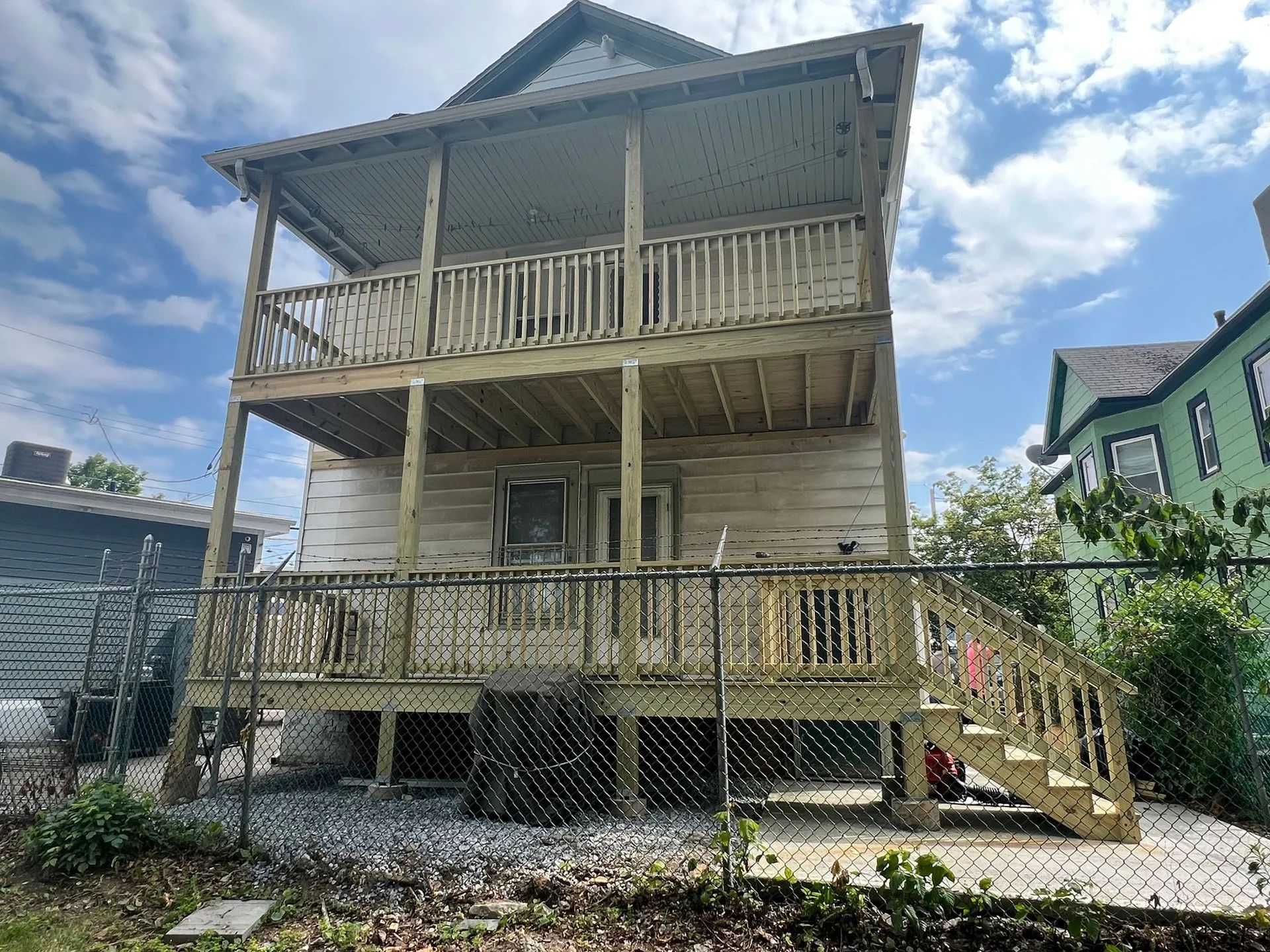 Back of a two-story weathered house with wooden decks and stairs, surrounded by a chain link fence and a cloudy sky.
