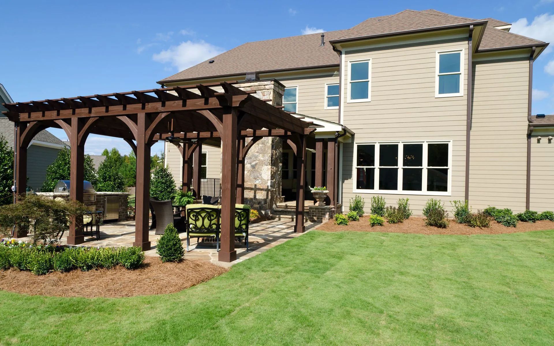 Brown pergola on a stone patio next to a beige house with a green lawn and blue sky.