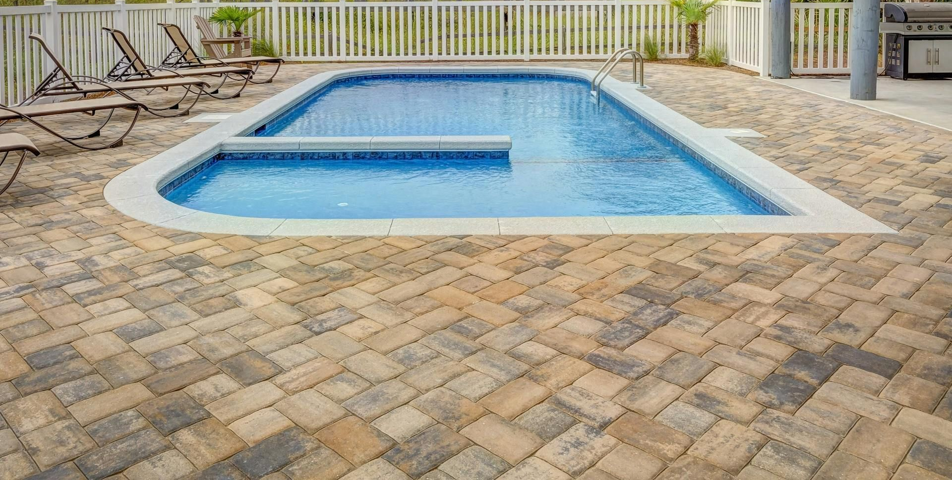 Swimming pool with a brick patio, lounge chairs, and a white fence.