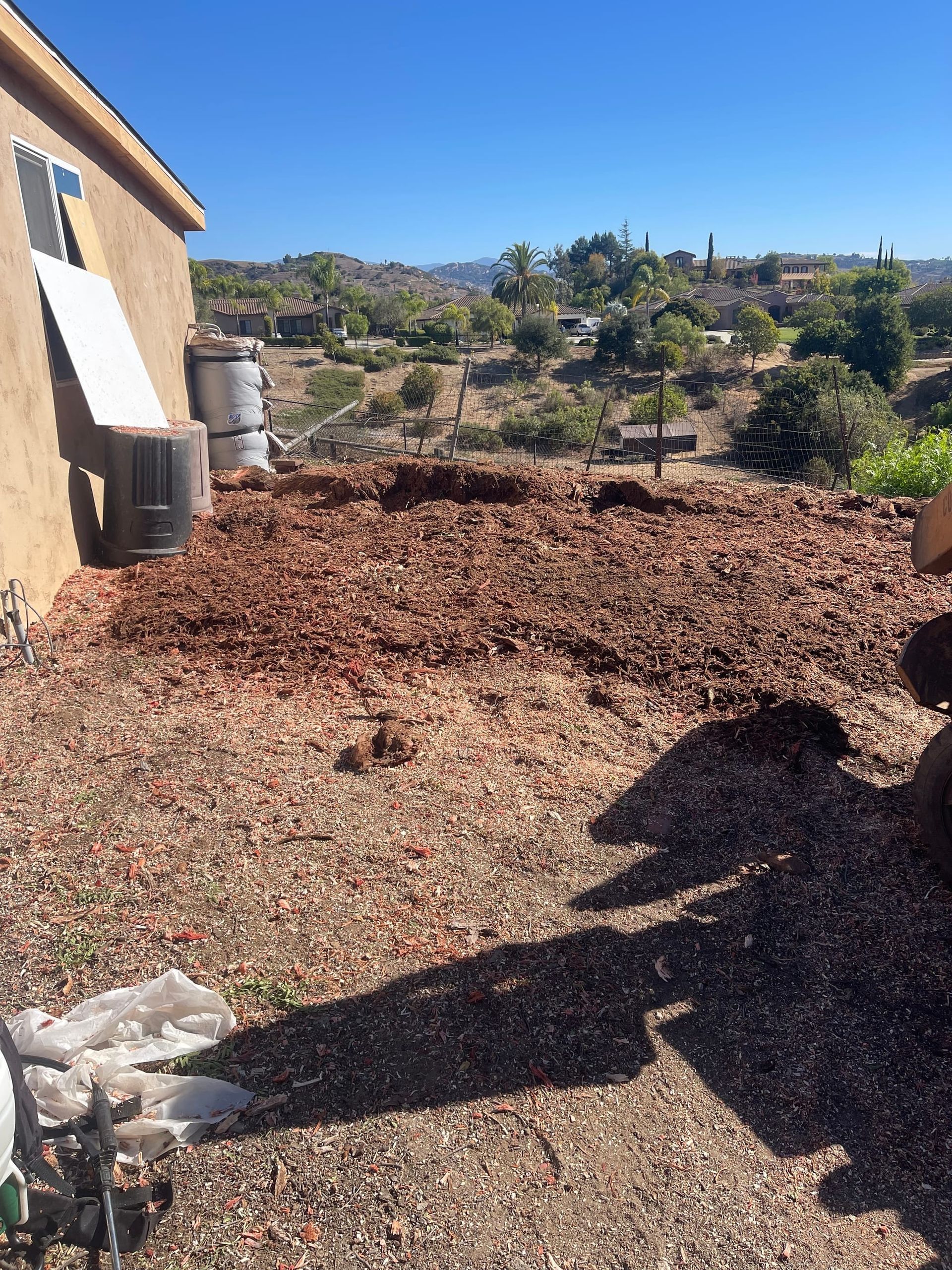Backyard with a tan house, mulch, and a hilly landscape under a clear blue sky.