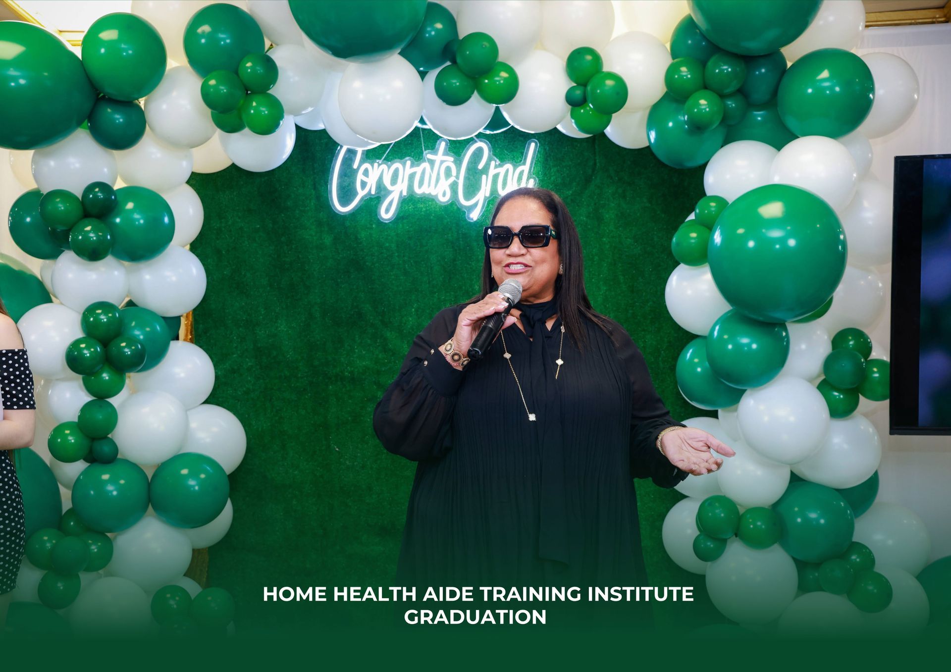 A woman is standing in front of a green and white balloon arch holding a microphone.