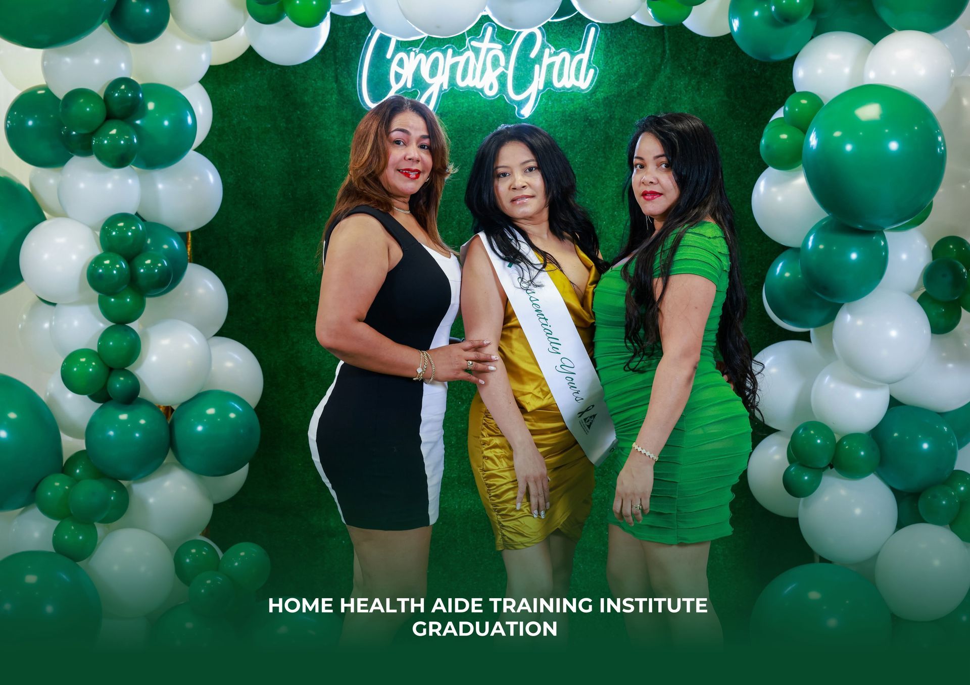 Three women are posing for a picture in front of a wall of green and white balloons.