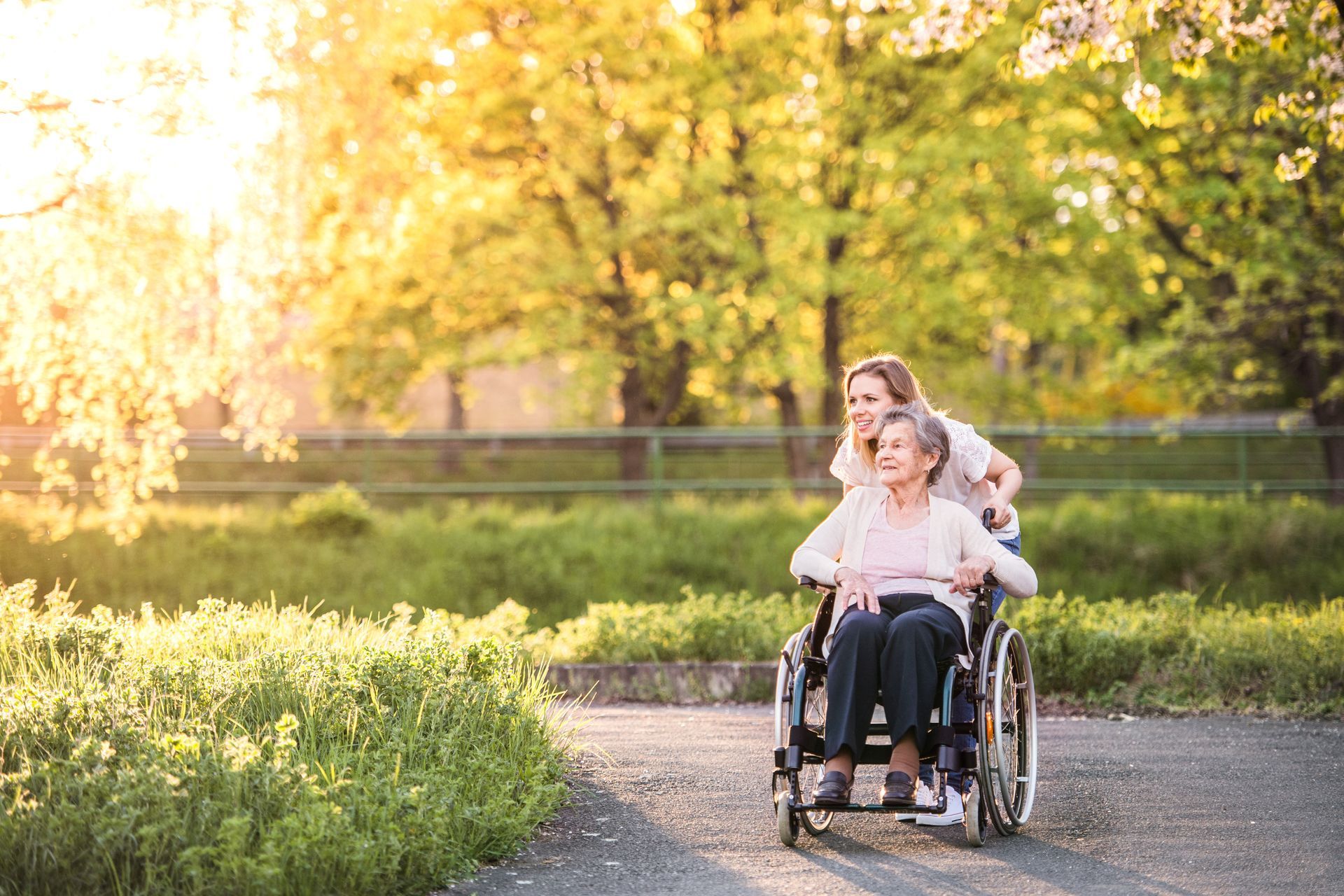 A woman pushes a person sitting in a wheelchair along a paved path in a sunlit park with trees and greenery.