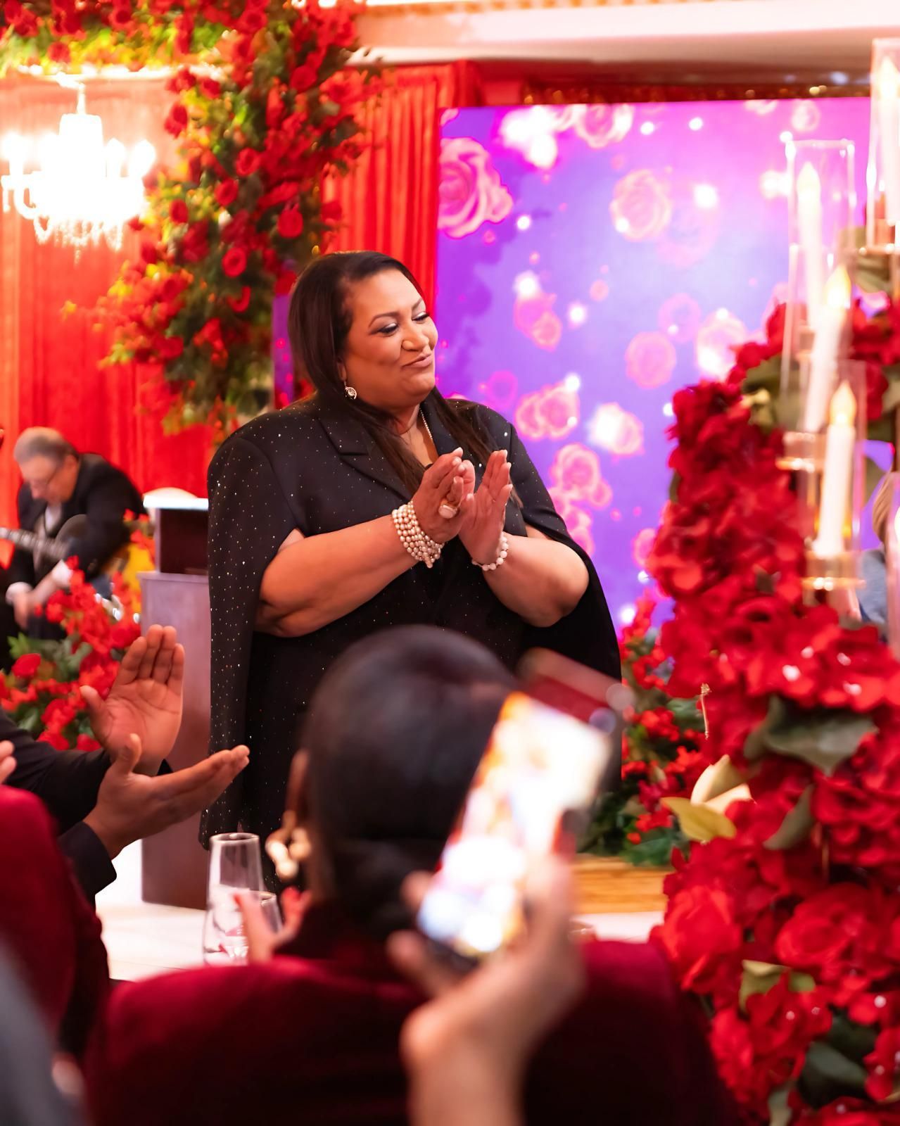 A woman is standing in front of a crowd of people in a room with red flowers.