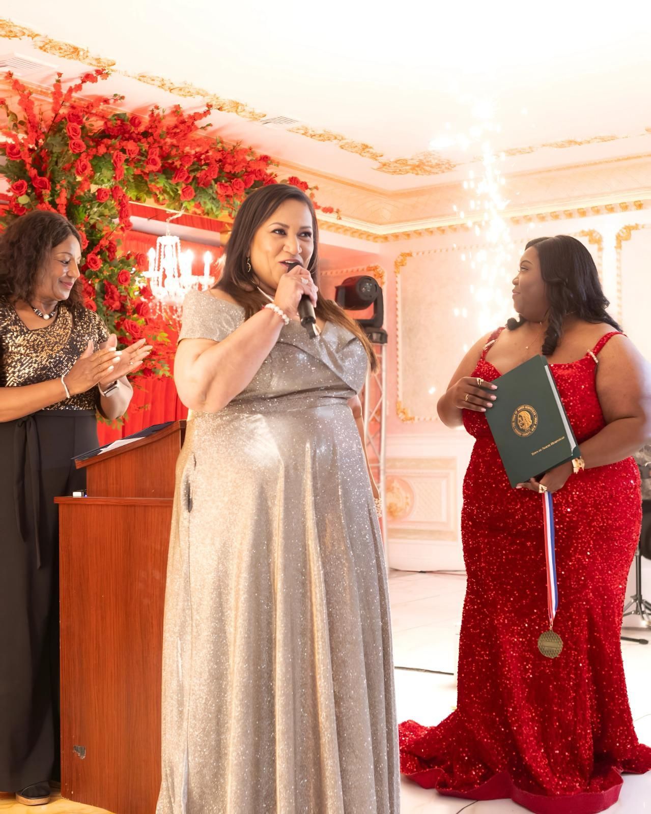 A woman in a silver dress is speaking into a microphone.