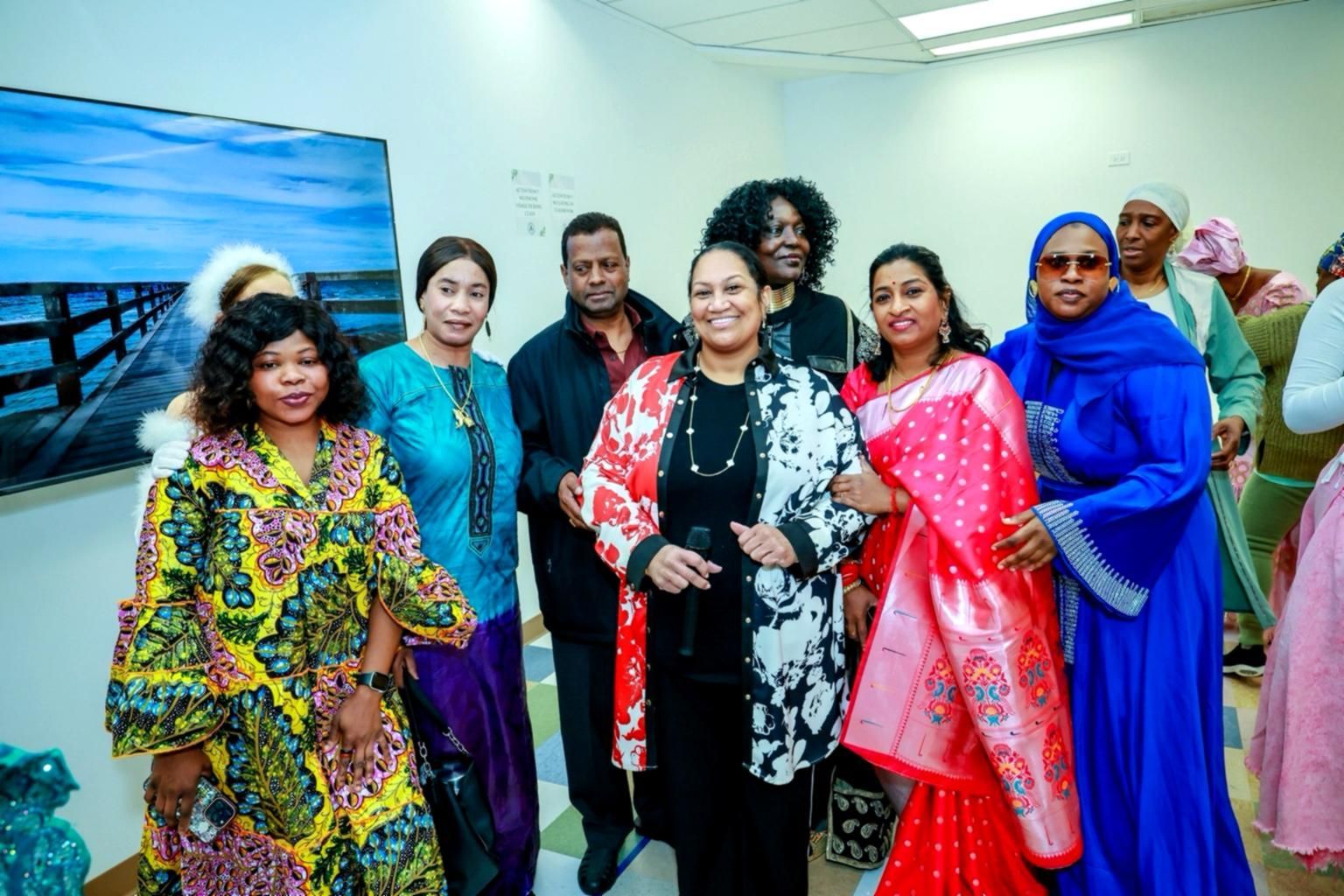 A group of people standing together indoors, some smiling, with a framed ocean landscape on the wall behind them.