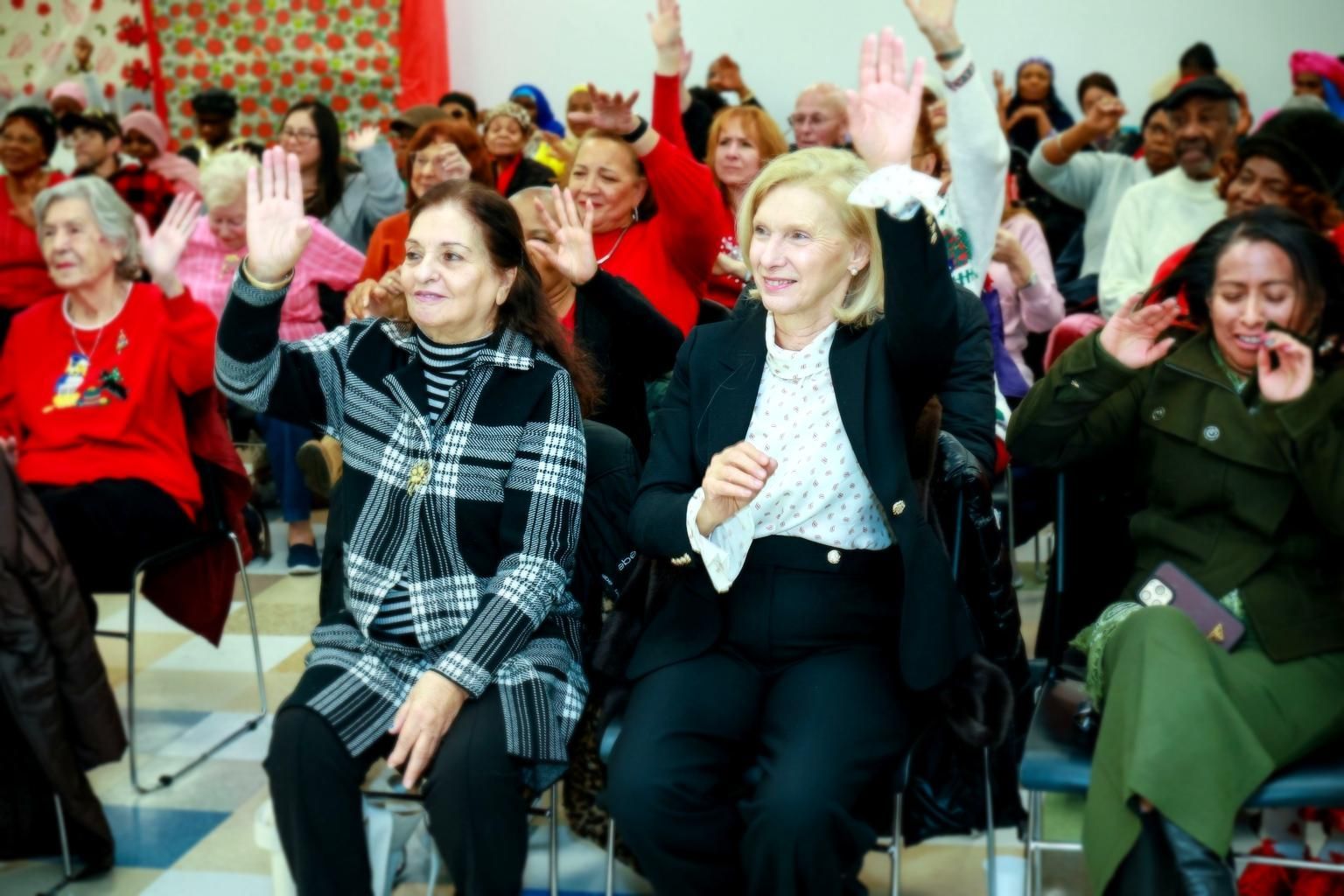 A diverse group of people sitting in a room, some with hands raised, during what appears to be a meeting or presentation.