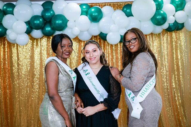 Three people stand side-by-side in front of a gold sequin curtain and a balloon arch, wearing sashes and smiling.