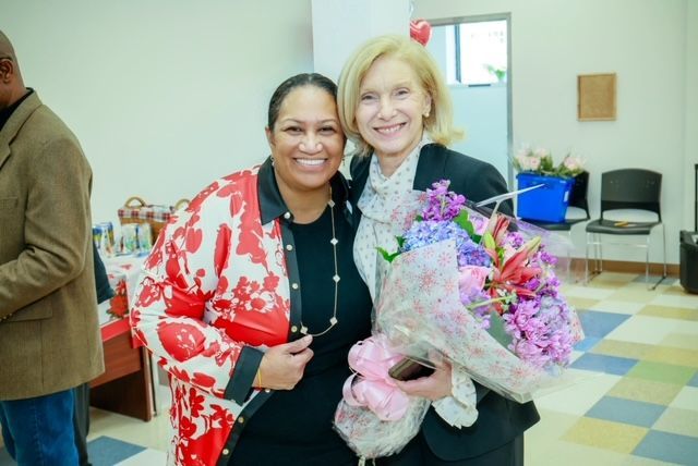 Two people pose smiling in an office setting; the person on the right holds a large bouquet of colorful flowers.