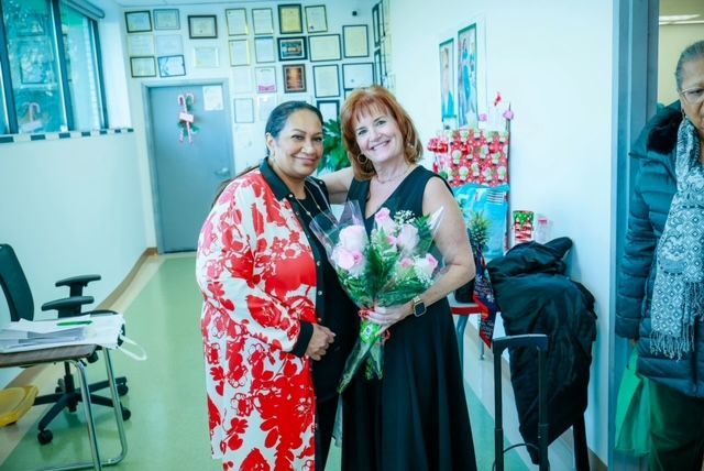 Two people smile while holding a bouquet of flowers in an office with framed documents on the wall.