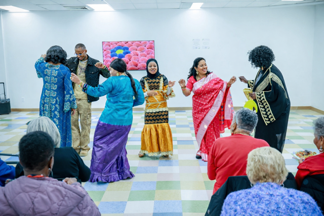 A group of people dance in a community room with a colorful patterned floor and a pink floral art piece on the wall.