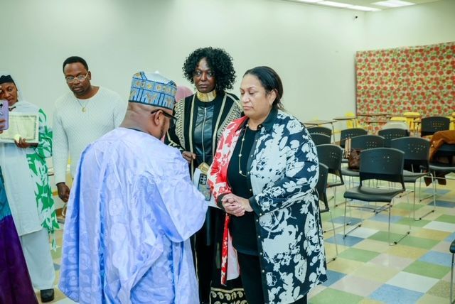 A person in a light blue traditional robe presents a plaque to a woman in a black and white patterned coat.