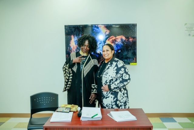 Two smiling people stand behind a desk with papers in a bright room, positioned in front of a digital screen.