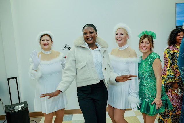 Four people stand indoors, including two in white festive outfits, one in a jacket, and one in a bright green dress.