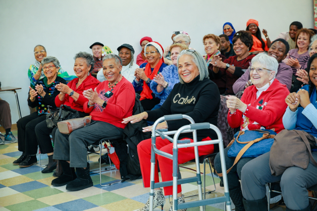 A diverse group of people sitting indoors, smiling and clapping, with one individual using a mobility walker in the front.