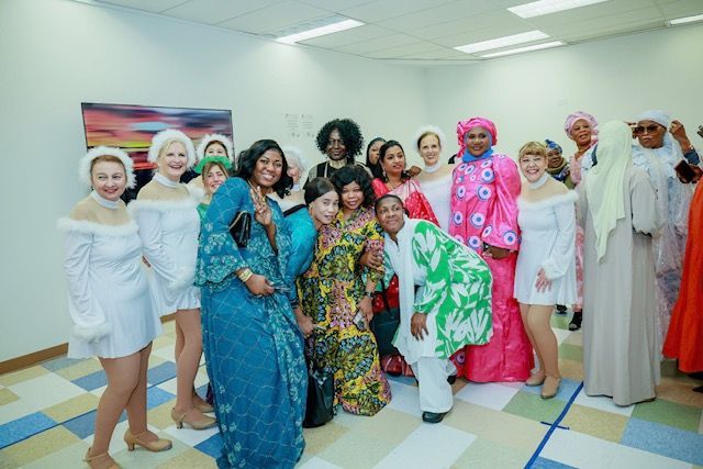A large group of people in diverse formal and festive attire pose for a photo in a bright room with tiled flooring.