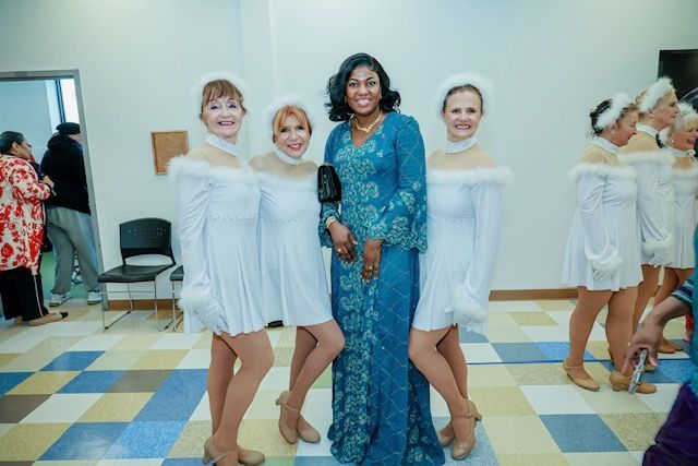 Three people in white, fur-trimmed dresses pose with a person in a long, teal lace gown in a room with a tiled floor.