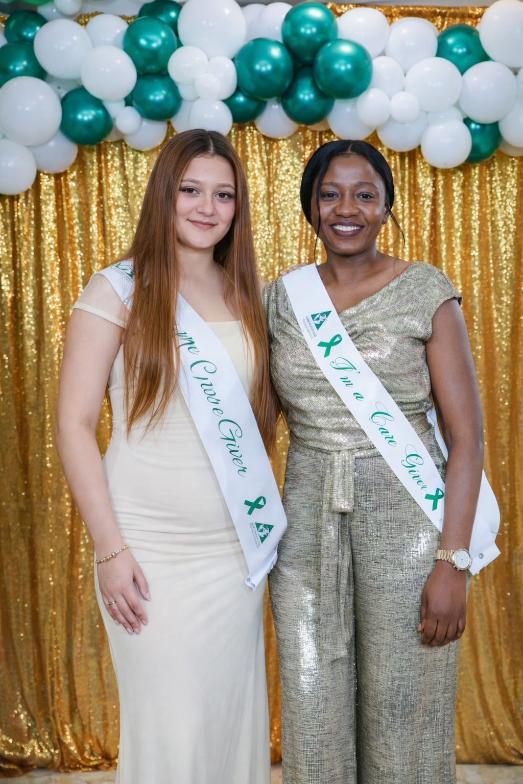 Two people in formal attire and pageant sashes stand smiling in front of a gold sequin backdrop and green and white balloons.