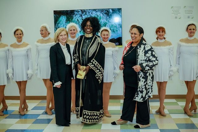 Two women in business attire stand centered before a row of performers in white dresses, posing for a group photo.