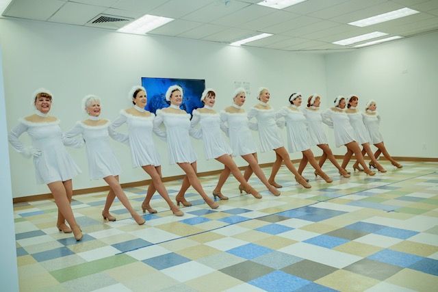 A line of dancers in white dresses and headbands performing a high-kick routine in a brightly lit studio.