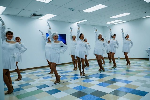 A group of dancers in white dresses and gloves pose in a bright, tiled room with one arm raised.