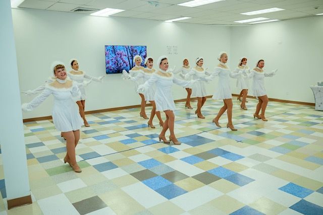 Dancers in short white dresses and headbands perform in a brightly lit room with a blue and white checkered floor.