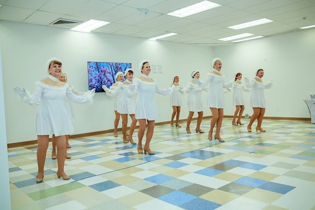 A group of performers in matching white dresses and headbands dance in a bright room with a blue and white tiled floor.