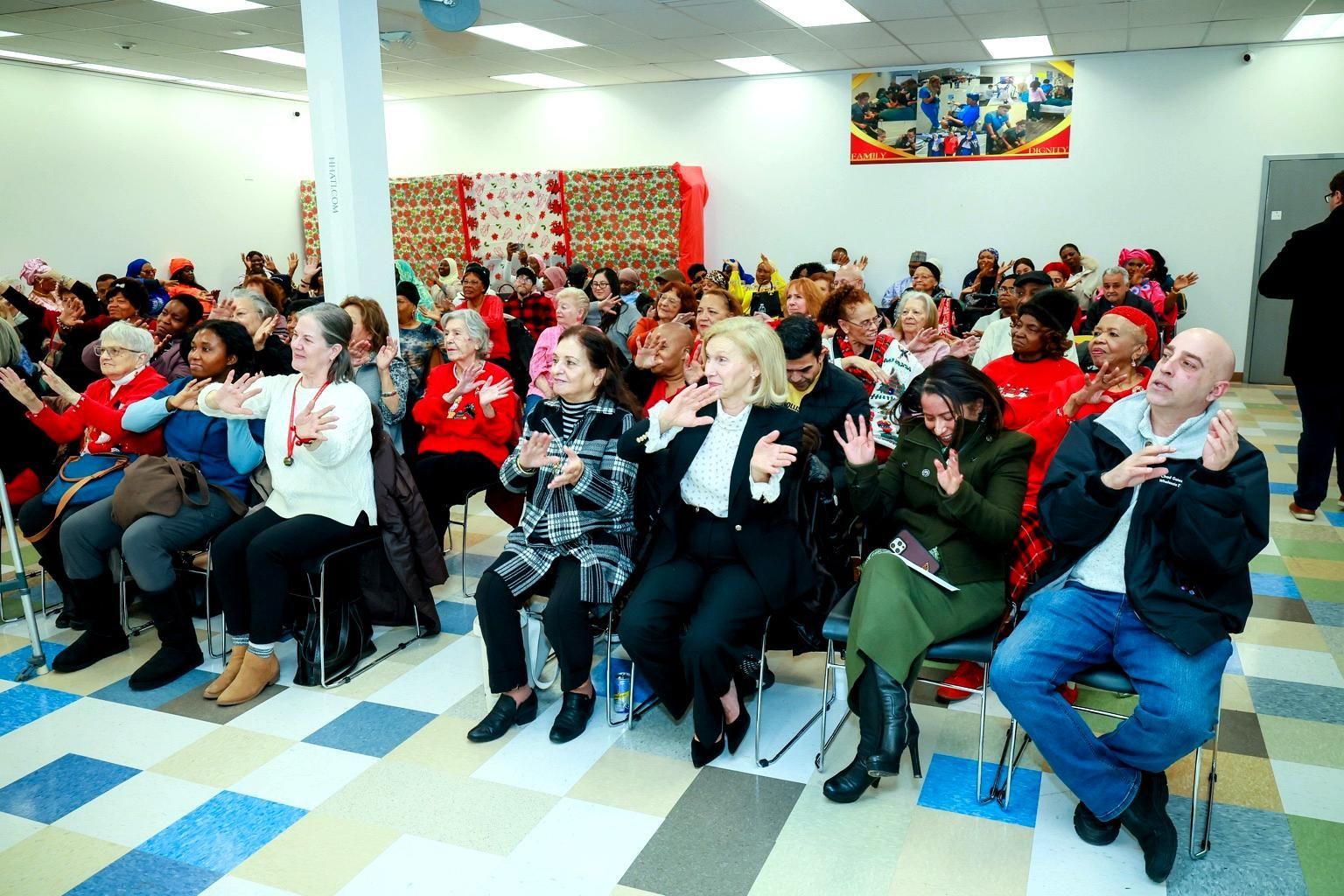 A group of people sitting in rows in a brightly lit hall, some clapping or smiling during an indoor event.