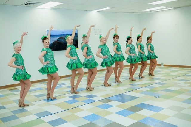 Eight dancers in matching green sequined dresses posing in a line in a bright studio with a tiled floor.